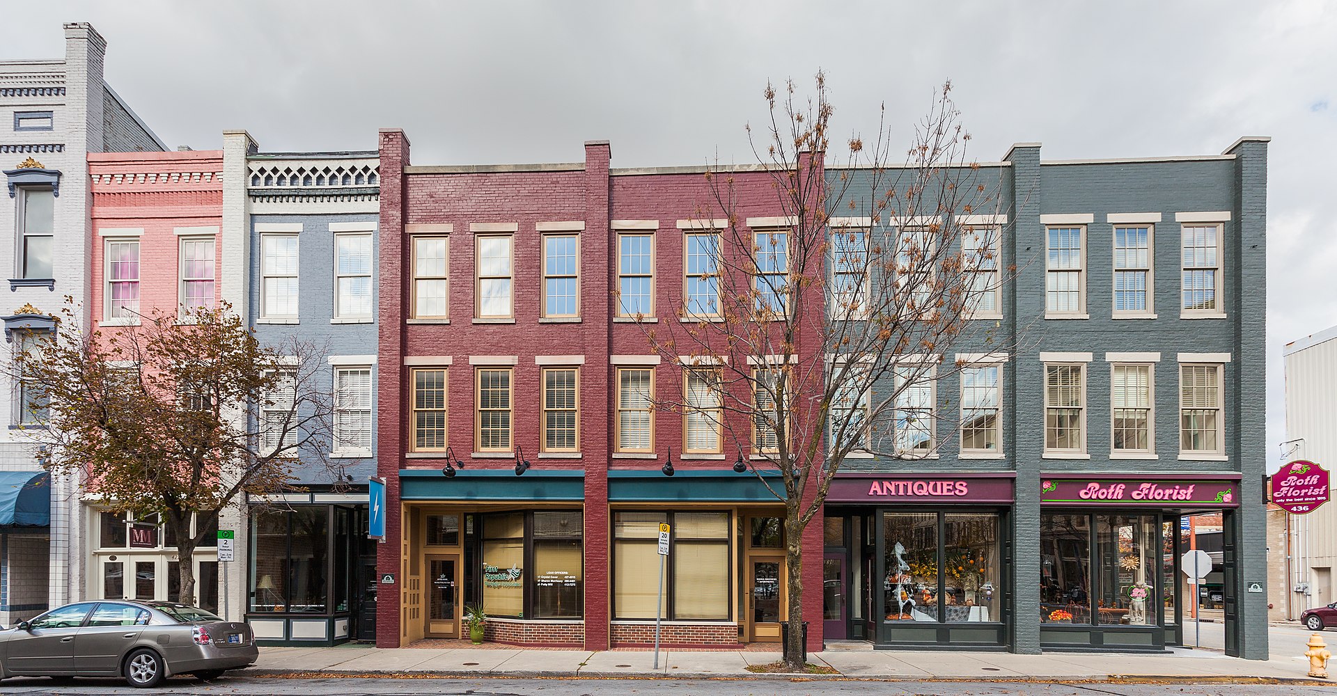 Row of three-story historic brick buildings with shops at street level, including an antiques store and a florist; parked car and leafless tree in front.
