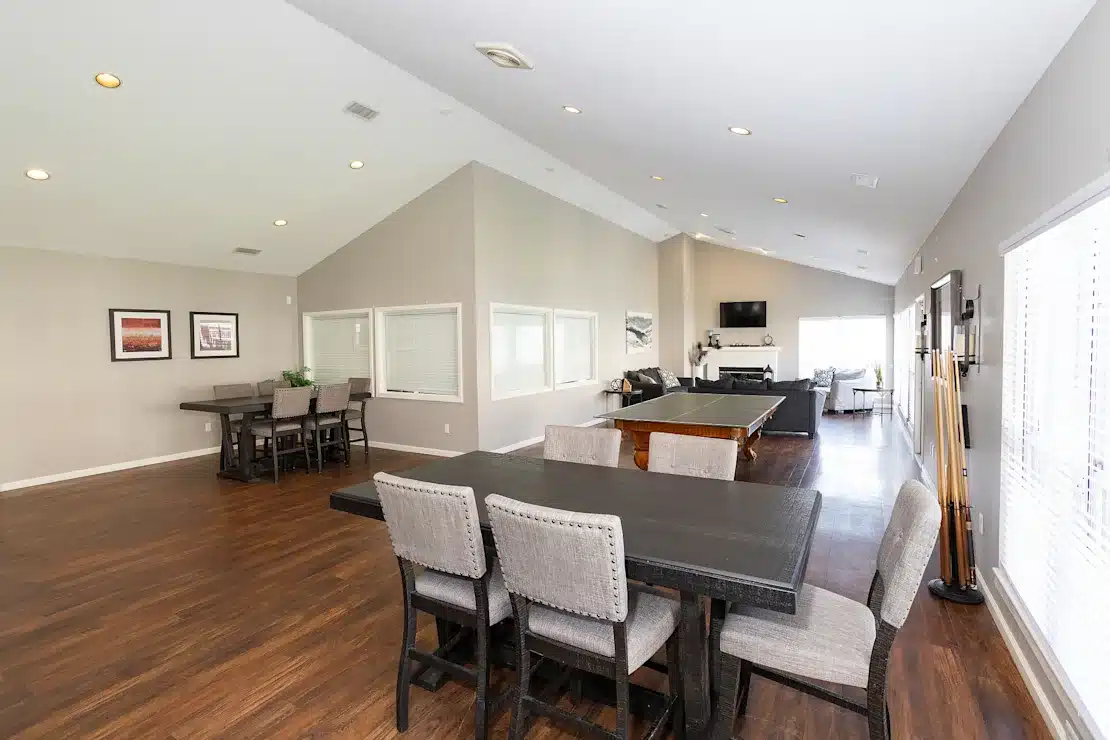 A spacious game room with hardwood floors, pool table, large TV, two dining tables with chairs, wall art, and windows letting in natural light.