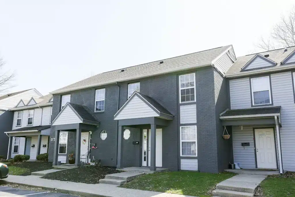 A row of two-story townhouses with gray siding, white trim, covered entryways, and small front lawns on a clear day.