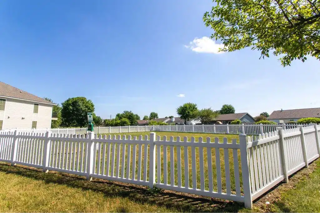 A white picket fence surrounds a grassy yard in a suburban neighborhood on a clear, sunny day.