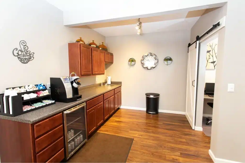 Modern kitchenette with wood cabinets, a coffee maker, snack station, and mini fridge. Wall decor and a sliding barn door are visible, along with a trash bin on hardwood flooring.