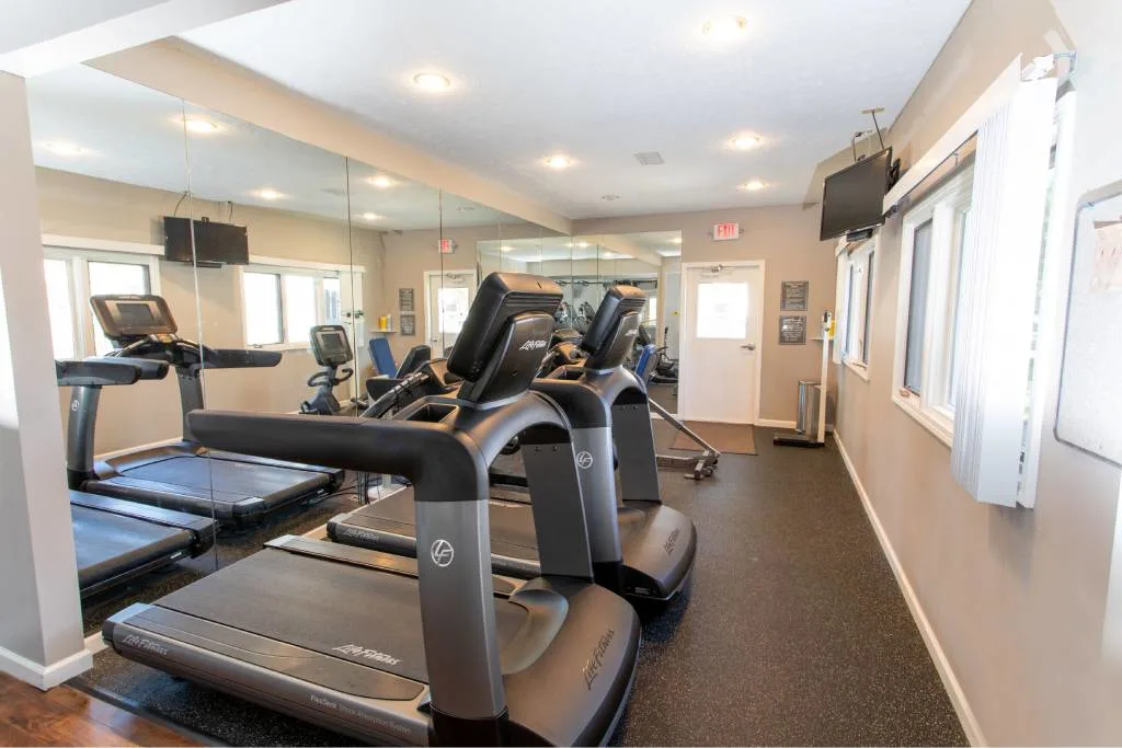 A brightly lit gym room with multiple treadmills facing wall mirrors, windows, and a mounted TV, featuring rubber flooring and neutral-colored walls.